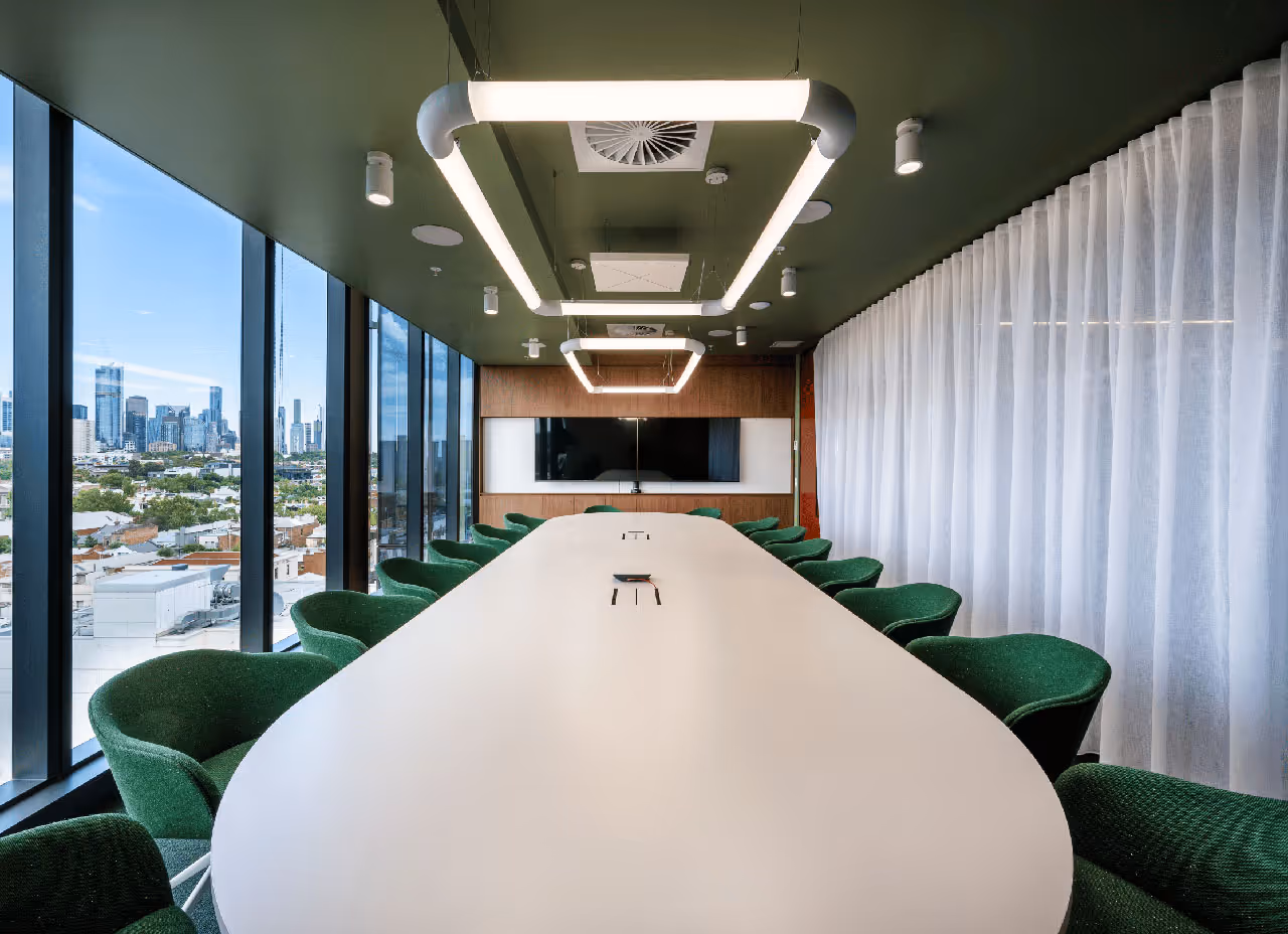 Modern conference room with long white table, green chairs, large windows showing city skyline, and suspended rectangular ceiling lights.