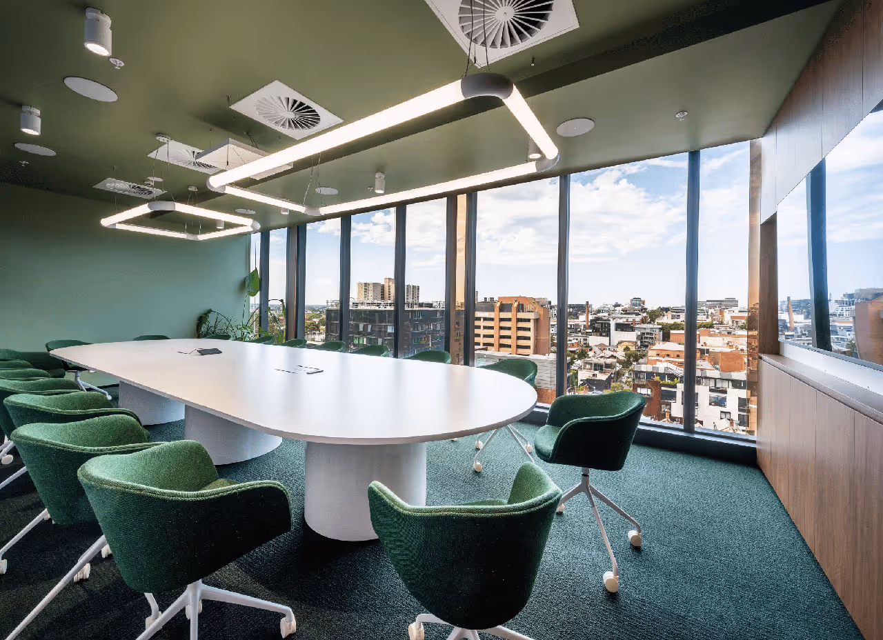 Modern conference room with large white oval table, green swivel chairs, hanging rectangular lights, and floor-to-ceiling windows showing cityscape.