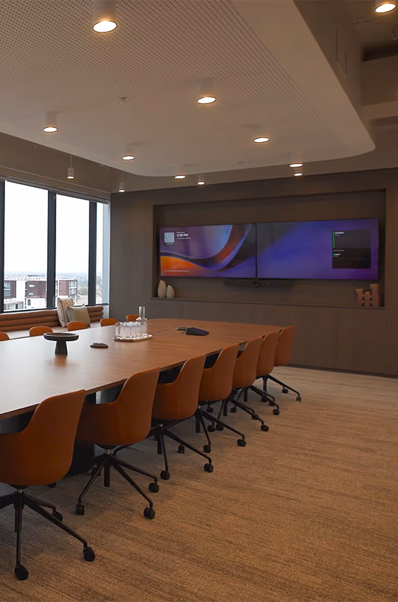 Modern conference room with a long wooden table, orange swivel chairs, a large wall-mounted dual screen, and large windows overlooking a cityscape.