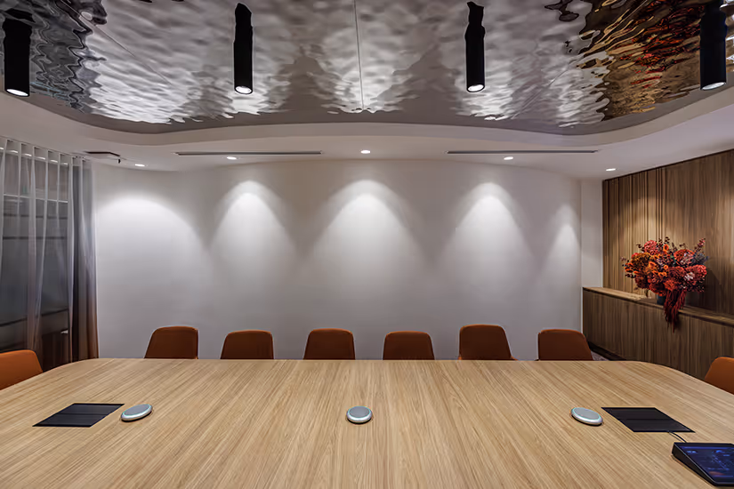 Modern conference room for Key Pharmaceuticals with a large wooden table, eight orange chairs, subtle wall spotlights, and a floral arrangement on a wood-panelled cabinet.
