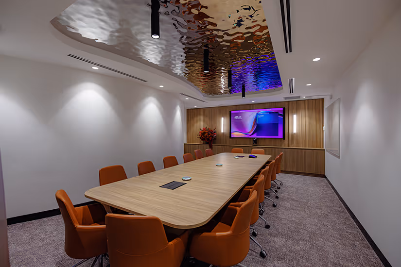 Modern boardroom for Key Pharmaceuticals with a long wooden table surrounded by orange chairs, a large screen on a wood-panelled wall, and a reflective textured ceiling panel.