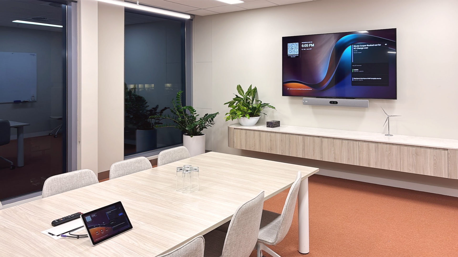 Modern conference room with beige chairs around a light wood table, wall-mounted TV displaying meeting info, and potted plants near the window.