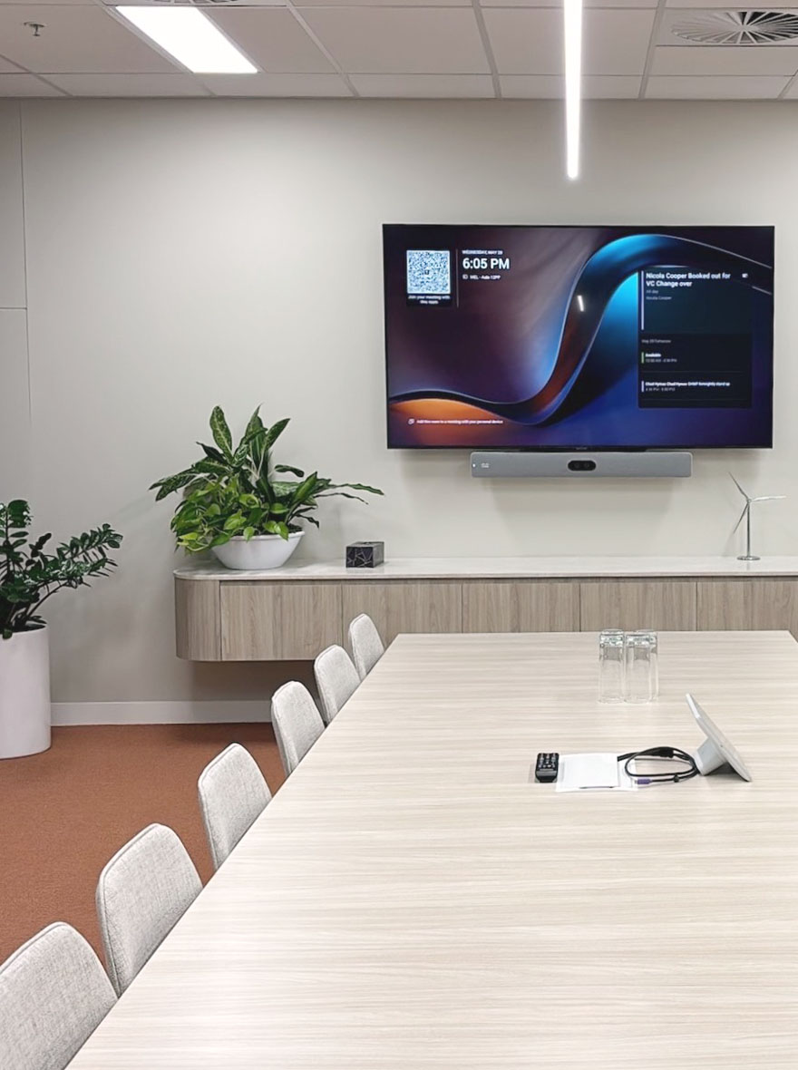 Modern conference room with a long light wood table, beige chairs, large wall-mounted screen displaying meeting details, and green plants on a sideboard.