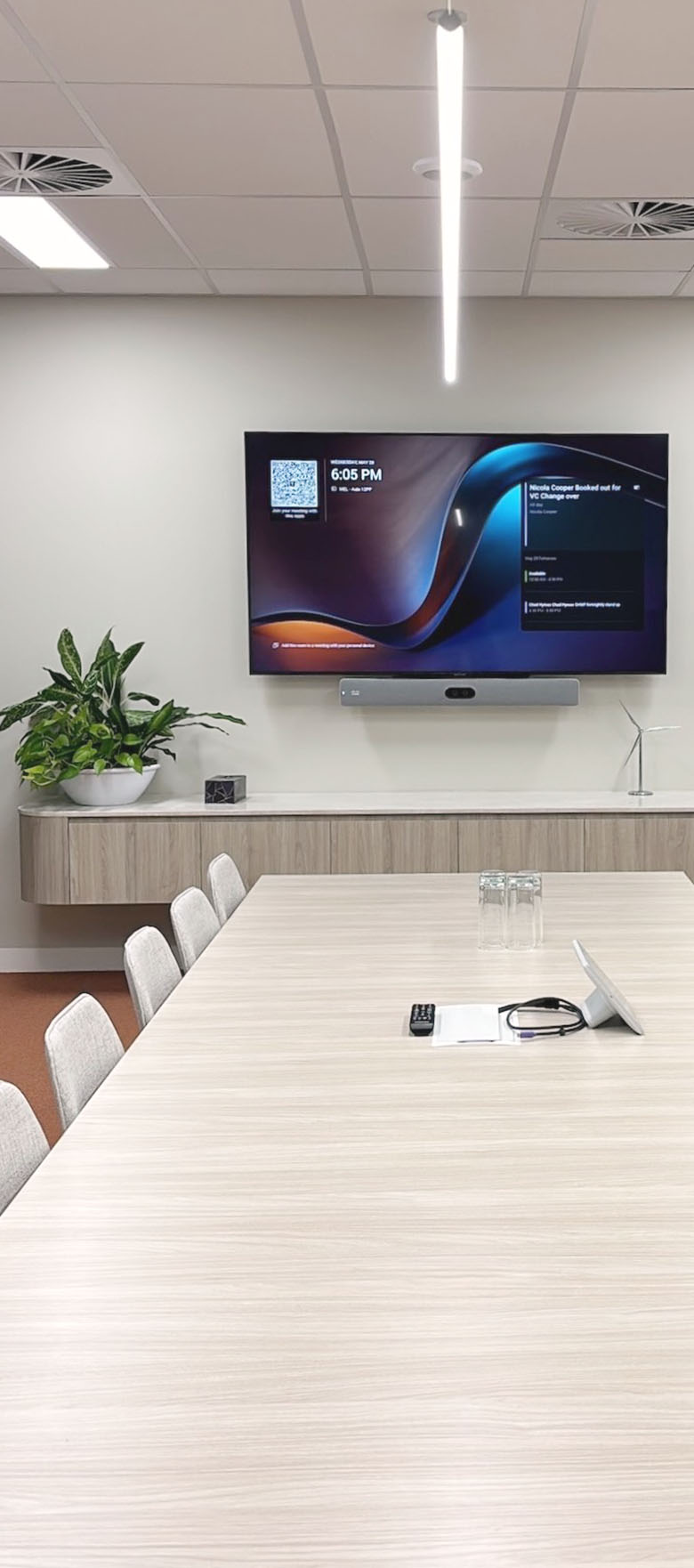 Modern conference room with a long light wood table, beige chairs, large wall-mounted screen displaying meeting details, and green plants on a sideboard.