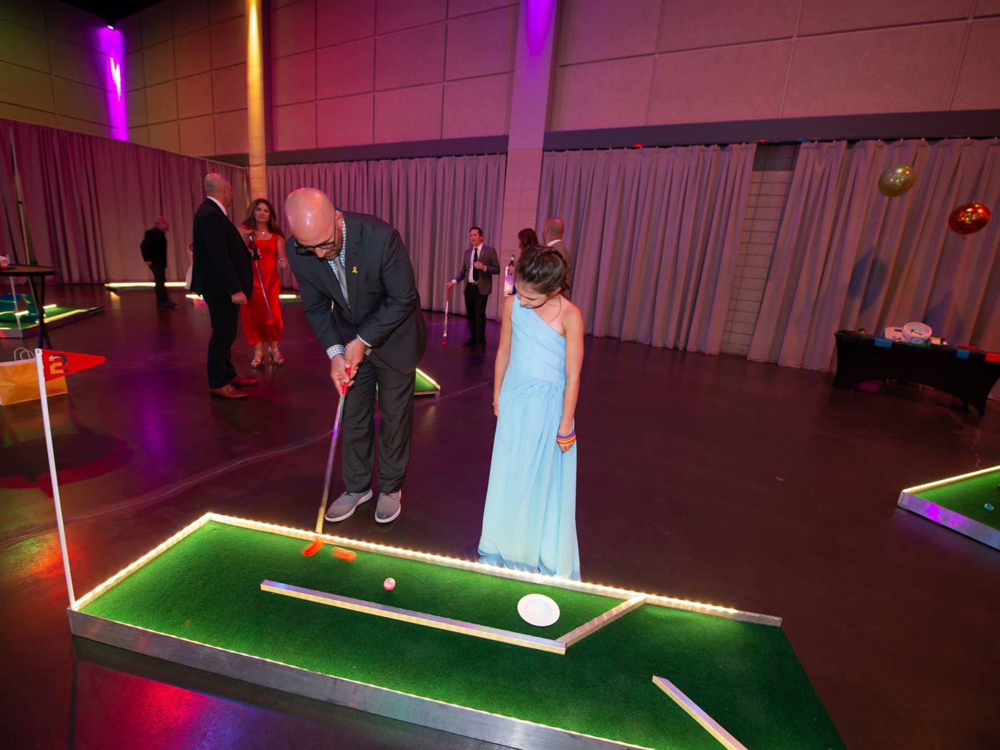 Wedding guest in a suit putting beside a girl in a blue gown on a lit mini golf hole at an evening reception