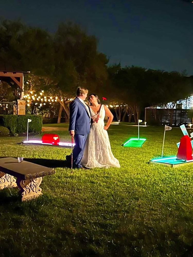 Bride and groom holding putters beside glowing mini golf holes at a night wedding reception