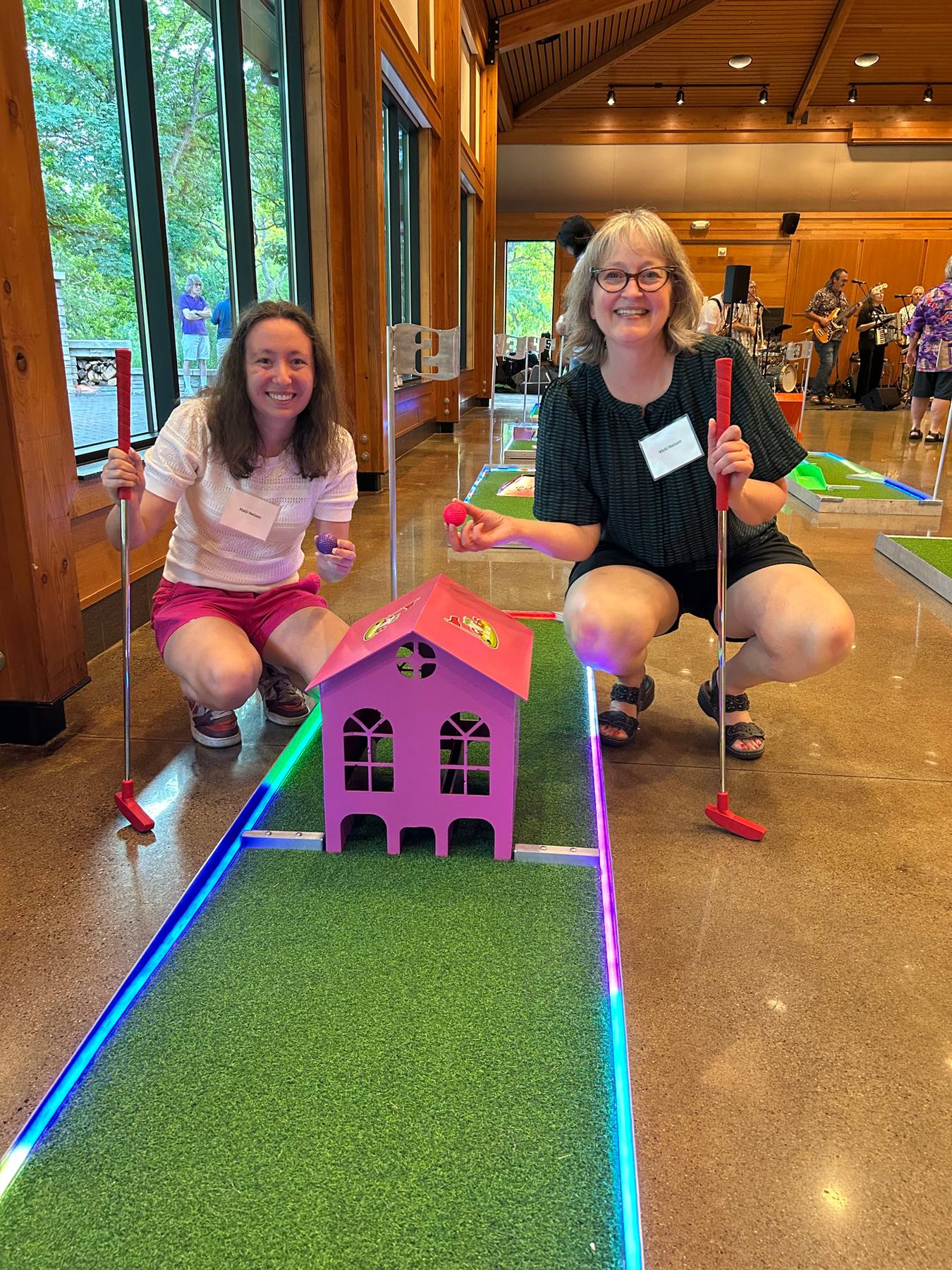 Two guests smiling beside a mini golf hole at a daytime event in a lodge