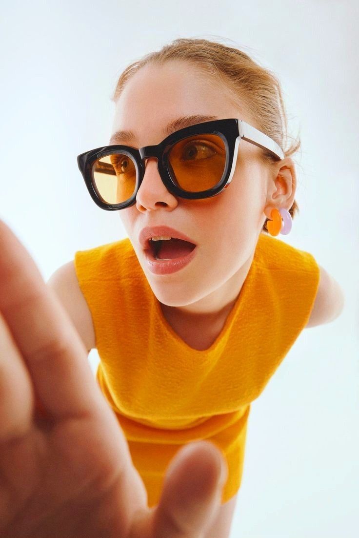 Fashion-forward woman wearing oversized black and white sunglasses with amber lenses, bold orange sleeveless dress, and colorful earrings, captured in a dynamic close-up pose against a minimalistic background.