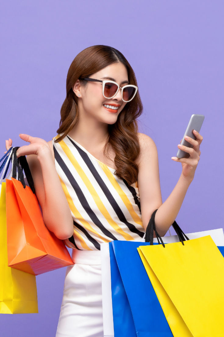Stylish woman wearing sunglasses and a striped top, smiling while holding colorful shopping bags and checking her smartphone, standing against a vibrant purple background, representing online shopping and retail fashion trends.