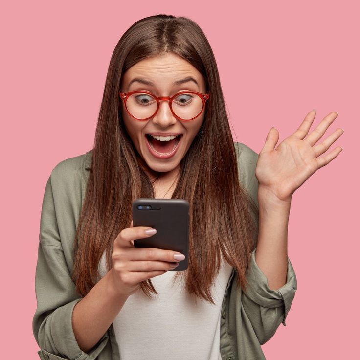 Excited young woman wearing red glasses and casual outfit, surprised while looking at her smartphone, isolated on a pink background, expressing joy and engagement with digital content.