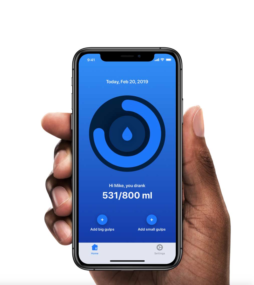 A person holding a smartphone displaying a water-tracking app. The app interface has a blue gradient background with a circular progress indicator showing water intake. The screen shows the date “Today, Feb 20, 2019,” and a message, “Hi Mike, you drank 531/800 ml.” Below the progress ring, there are two buttons labeled “Add big gulps” and “Add small gulps.” The navigation bar at the bottom has icons for “Home” and “Settings.” The phone has a black frame with a notch at the top, showing the time “9:41” and battery, Wi-Fi, and signal indicators.