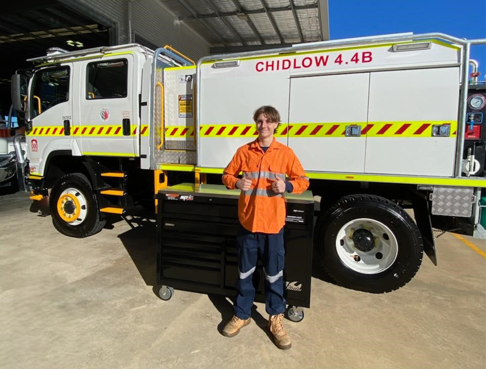 A student stood in hi-vis gear in front of a tool-box and truck