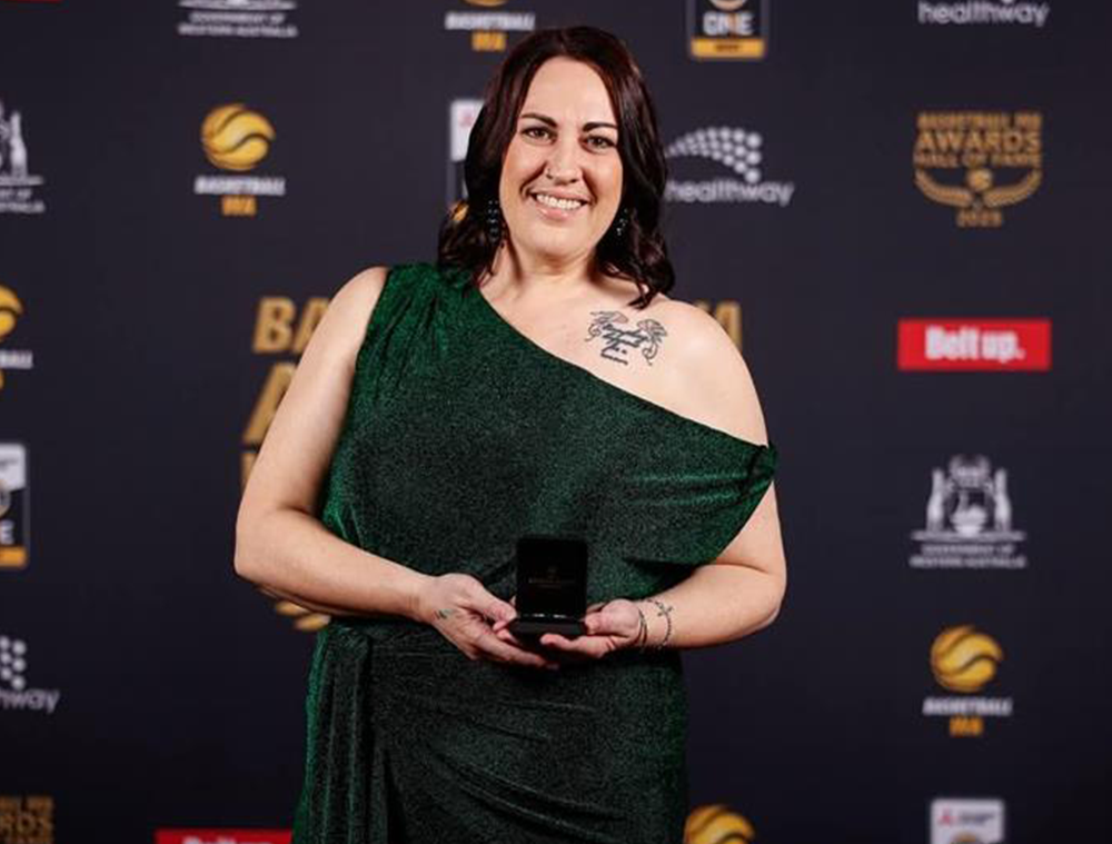 A woman smiling and standing in front of a branded media wall at an awards event holding a small open box with an award inside