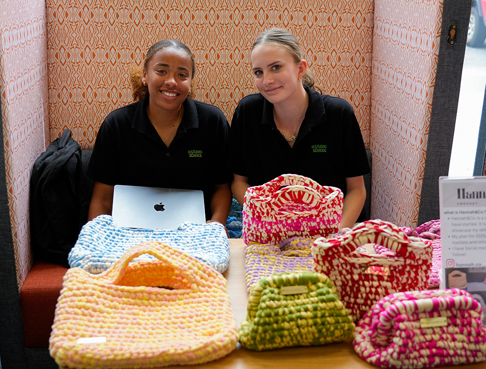 Two female students sat behind a table full of crochet bags