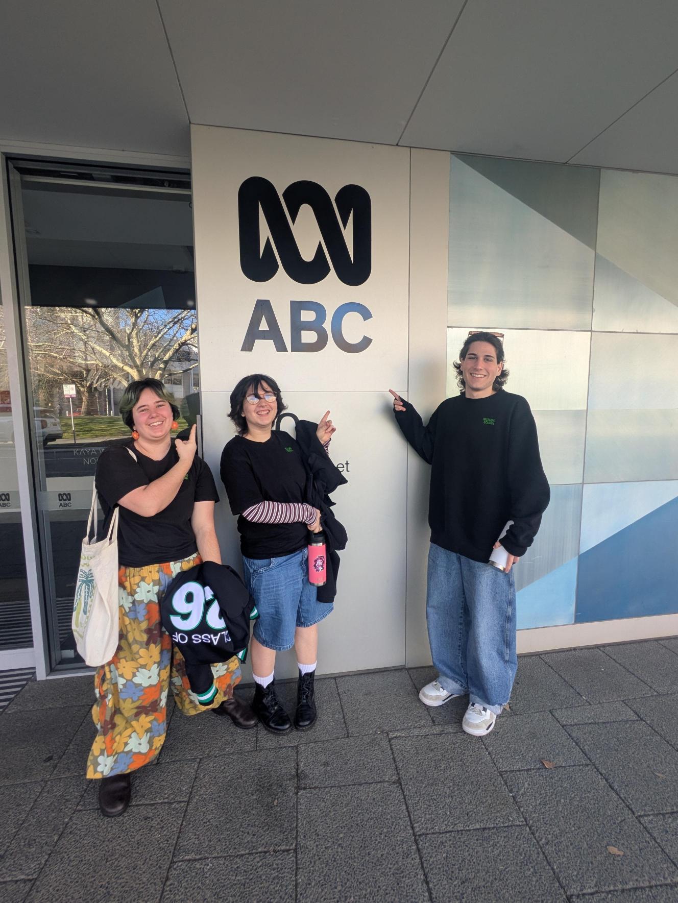 Three students stood outside the ABC Radio station building pointing at the logo