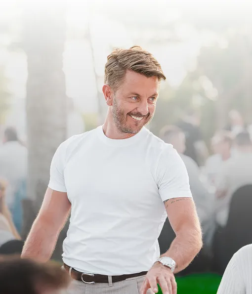 Ein lächelnder Matthias Aumann mit kurzem braunem Haar trägt ein weißes T-Shirt und eine Armbanduhr, steht im Freien bei Tageslicht, umgeben von verschwommenen Menschen im Hintergrund.