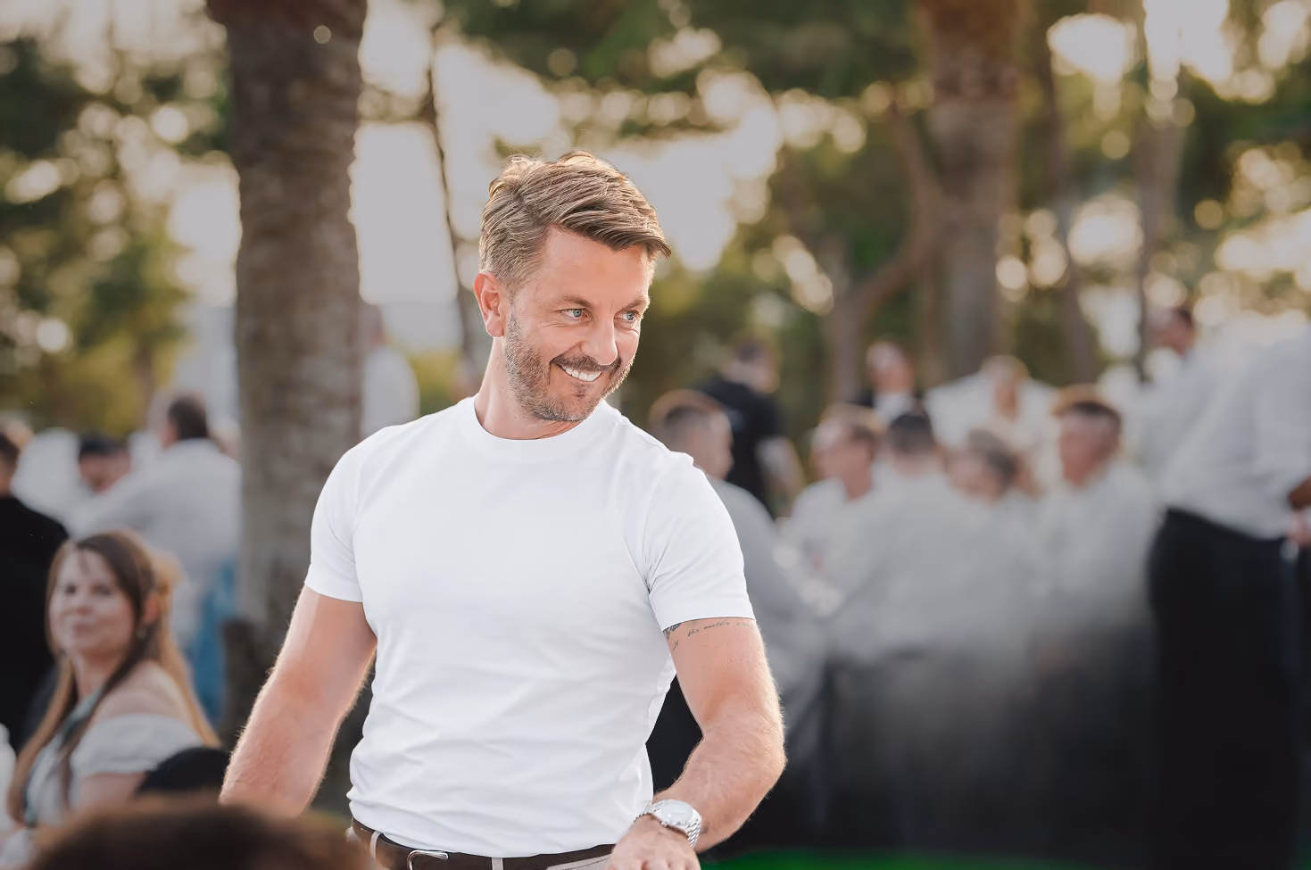 Ein lächelnder Matthias Aumann in weißem T-Shirt mit Armbanduhr steht im Freien vor unscharfen Menschen und Bäumen im Hintergrund.