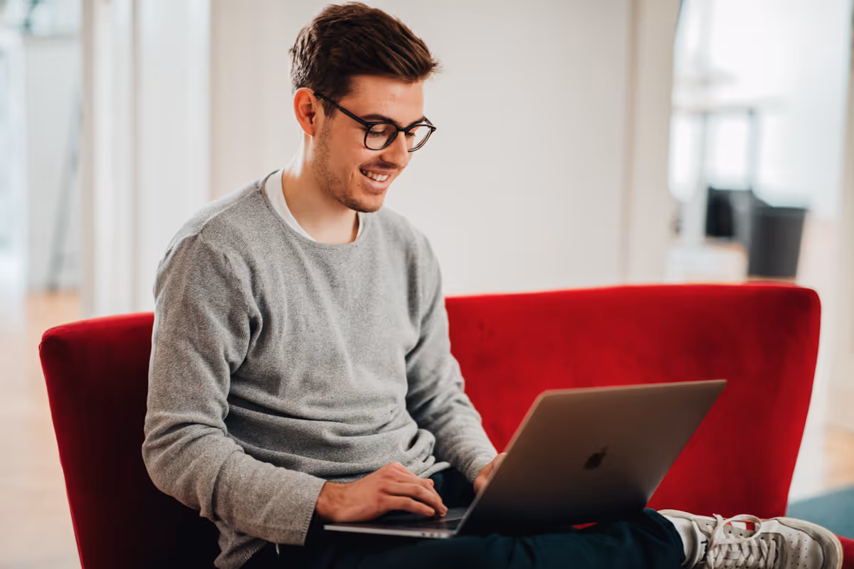 Agentur-Mitarbeiter sitzt auf einem Sofa und arbeitet mit seinem Macbook auf dem Schoß.