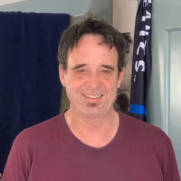 Smiling man with dark hair wearing a maroon T-shirt standing indoors with towels and a door in the background.