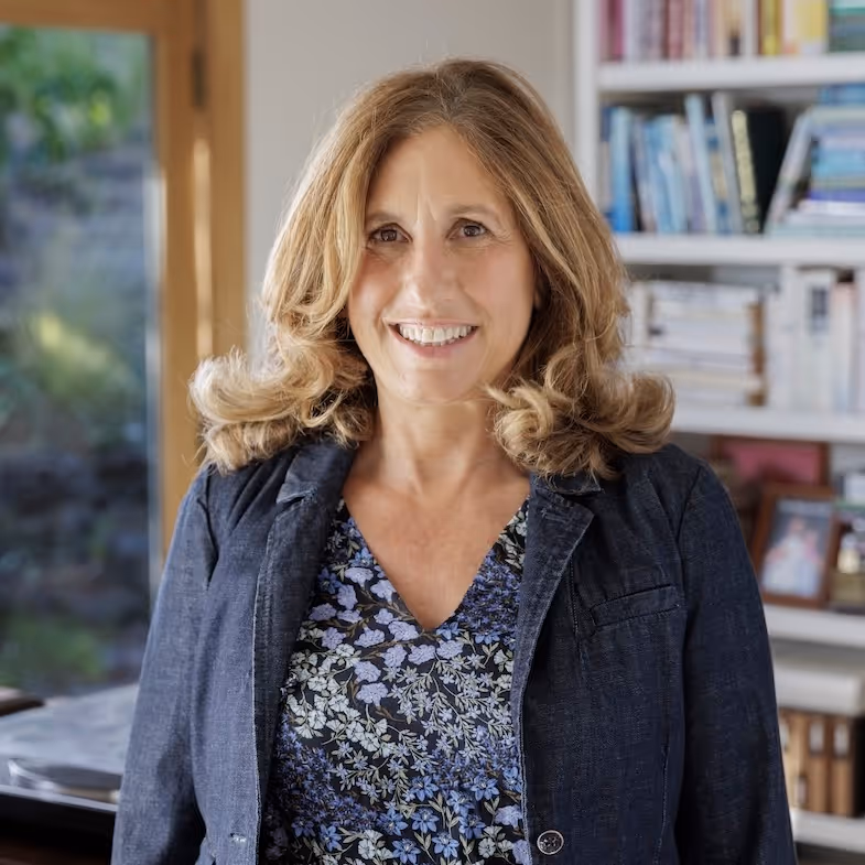 Woman with shoulder-length wavy blonde hair wearing a floral blouse and dark blazer standing in a room with bookshelves and a window.