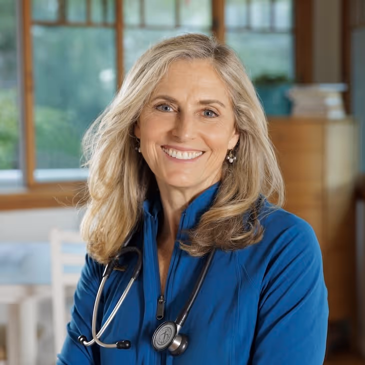 Smiling female healthcare professional with blonde hair wearing a blue jacket and stethoscope around her neck in a room with windows and wooden furniture.