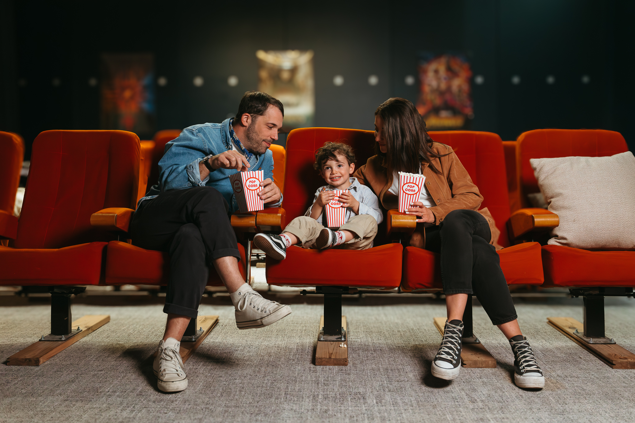 Une famille assise dans une salle de cinéma sur des fauteuils rouges, partageant du pop-corn et souriant ensemble.