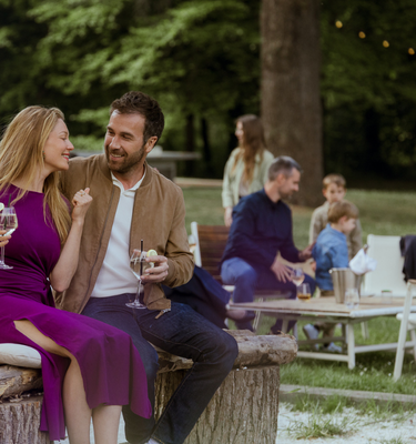 Couple seated outdoors, enjoying drinks and smiling at each other in a park setting with others in background.