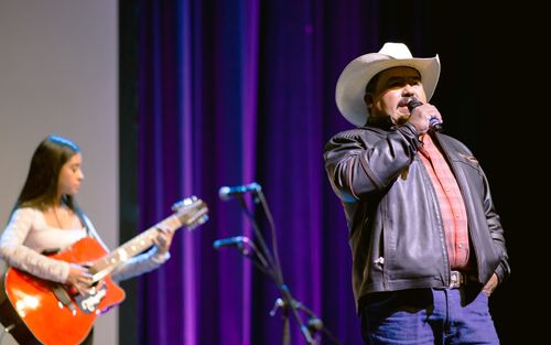 Rodolfo Carranza, a man in a tan cowboy hat and dark leather jacket, sings on a stage. Behind him, Saira Aldaco, a young woman in a white, long-sleeved shirt, plays guitar.
