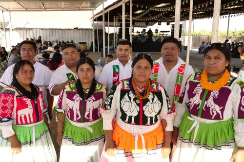 A group of dancers faces the camera; there are four women standing in front of four men. The women all wear white dresses with colorful, bright embroidery; the men wear white, button-down shirts which are also decoratively embroidered.
