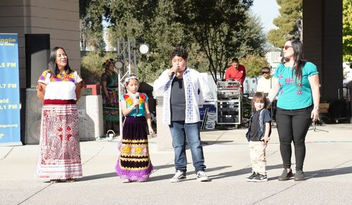 On a bright, sunny day, two women and three children sing in front of a building. Audio technicians are visible in the background.