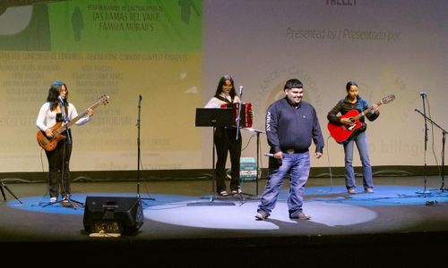 A distance photo of Adrián Sánchez Ávila, a young man in a dark blue, long-sleeved shirt, standing beneath a spotlight on a stage. Behind him are three young, women musicians.
