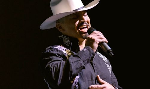 A dramatically lit, close up image of Félix Arreola singing. He wears a white hat and a dark, embroidered shirt. One of his hands holds a mic; the other rests on his chest.