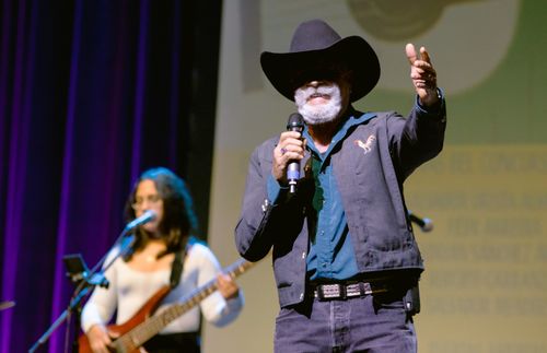 Salvador Mendoza, a man with a dark blue jacket, a cowboy hat and a bushy, white mustache and beard, sings. Behind him, a young, woman musician in a white shirt plays bass.