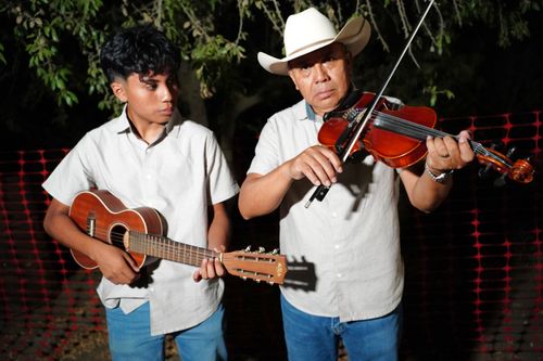 Two men, one younger and one older, pose for a photo at night. They both wear white, button-down shirts and blue jeans. The younger man plays guitar; the older, violin.