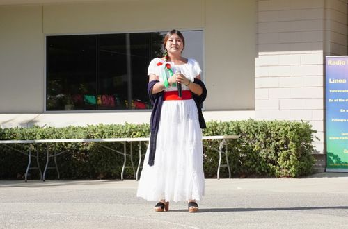 A woman stands outside a building holding a microphone in both of her hands. It is sunny, and she wears a long, white dress, a dark shawl around her elbows, a red sash around her waist, and green and red ribbons entwined in her braid.