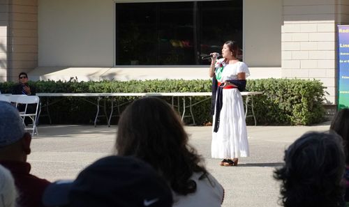 A distance photo of a woman in a long, white dress singing to a crowd in front of a building on a sunny day.