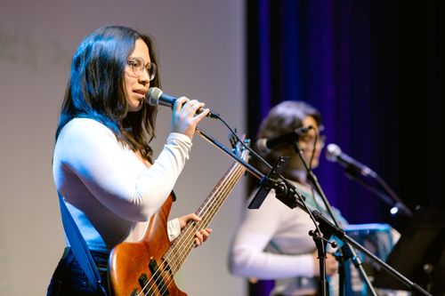 Two young women musicians in matching white, long-sleeved shirts perform on stage. The woman on the left is a bassist singing into a mic. The woman to her right is an accordionist.