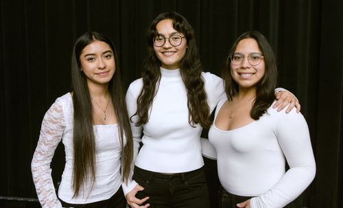 Three young women with dark hair, dark pants, and white, long-sleeved shirts stand close together and smile against a black background.