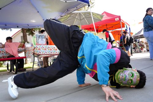Gabriel Gutierrez, whose whole face is covered by a decorative, green mask, performs a breakdance freeze. His head, hands, and left foot touch the ground, while his right leg is suspended in the air.