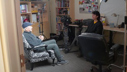 A photo taken from outside of a small room. Inside, a man with white hair and dark clothes sits on an armchair, facing a woman seated in the shadows.