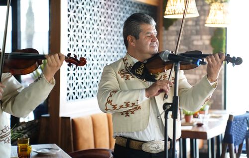A man in an off-white mariachi outfit closes his eyes as he plays violin in a restaurant.