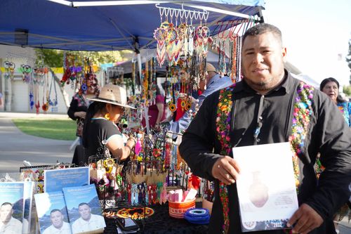 At a community festival, a man stands in front of his booth, which displays finely beaded artworks and jewelry.