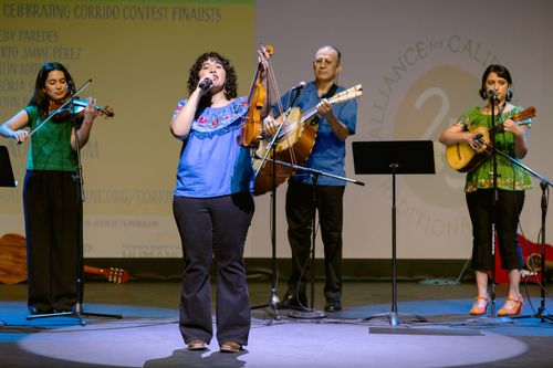 A woman in a blue shirt sings on stage. Behind her are three musicians: a woman playing violin, a man playing guitar, and another woman playing guitar.