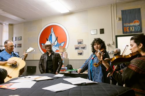 Four people gather around a covered piano, upon which lies scattered sheets of music. On the left, there are two men. One plays guitar, and one sings. On the right are two young women playing violin.