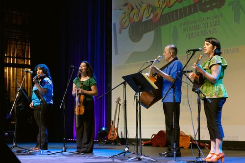 A distanced photo of four musicians on a stage. From the left, the first two are women playing violin, followed by a man playing guitar and a woman playing guitar.