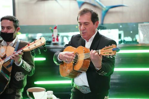 Two men, who are mariTwo men, who are mariachis dressed in black, perform in a restaurant. The one on the left plays a guitar, while the one on the right plays a vihuela.achis dressed in black, play guitar in a restaurant with fluorescent green lights.