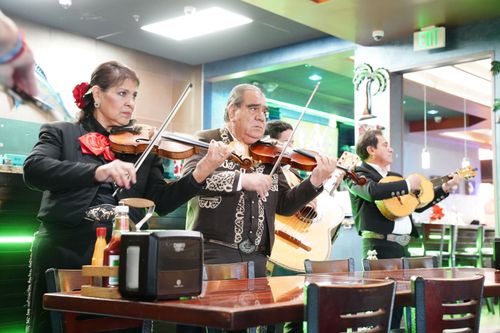 Four mariachis, dressed in black, perform in a brightly lit restaurant. Furthest left is a woman violinist; the other three musicians are men: a violinist, a guitarist, and vihuela player.