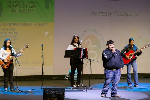 A distance photo of Adrián Sánchez Ávila, a young man in a dark blue, long-sleeved shirt, singing beneath a spotlight on a stage. Behind him are three young, women musicians.