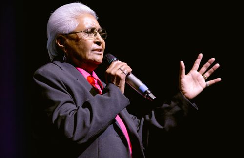 A close-up photo of a woman with short, white hair singing, one hand extended expressively in front of her.