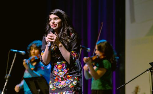 A close-up photo of a woman in a black dress, embroidered with colorful flowers, singing on a stage. Two women play the violin behind her.